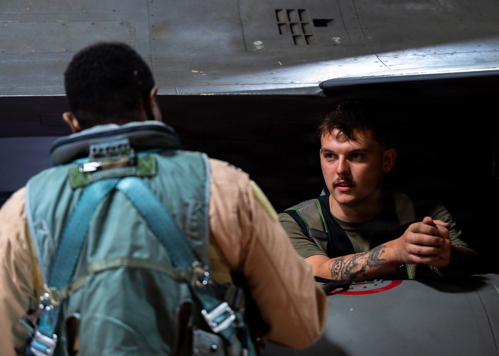 Two military personnel are interacting near a military aircraft, likely in a hangar or flight deck environment. One individual is seated in or leaning out of the aircraft cockpit area, appearing to communicate with the other person who is wearing flight gear including a harness and survival vest. The scene suggests a pre-flight or post-flight briefing or equipment check.
