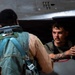 Two military personnel are interacting near a military aircraft, likely in a hangar or flight deck environment. One individual is seated in or leaning out of the aircraft cockpit area, appearing to communicate with the other person who is wearing flight gear including a harness and survival vest. The scene suggests a pre-flight or post-flight briefing or equipment check.