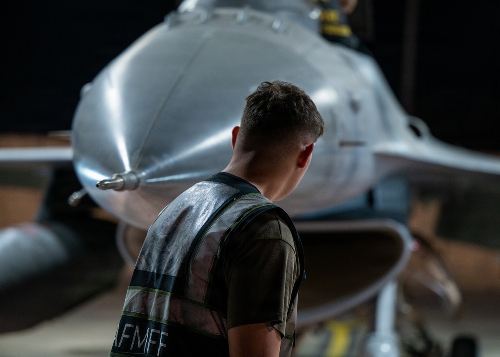 A military aircraft maintainer or ground crew member is inspecting the nose section of what appears to be an F-16 Fighting Falcon jet fighter in a hangar setting. The individual is wearing a reflective safety vest with partial text reading 'AFMFF' visible, suggesting US Air Force affiliation. The image is taken from behind the person, emphasizing the scale and polished metallic nose cone of the aircraft.