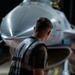 A military aircraft maintainer or ground crew member is inspecting the nose section of what appears to be an F-16 Fighting Falcon jet fighter in a hangar setting. The individual is wearing a reflective safety vest with partial text reading 'AFMFF' visible, suggesting US Air Force affiliation. The image is taken from behind the person, emphasizing the scale and polished metallic nose cone of the aircraft.