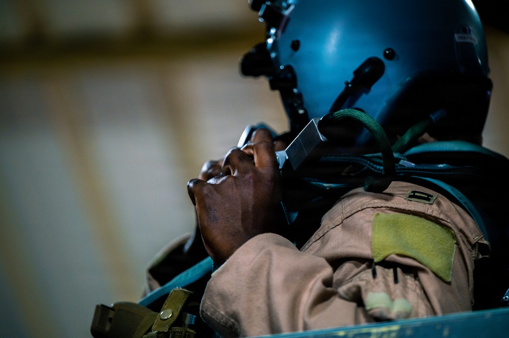 A military aviator or aircrew member is photographed in close-up profile view, wearing a flight helmet and flight suit. The individual is looking upward, with one hand raised near the chin area. The image is taken in low-light conditions with dramatic side lighting emphasizing facial features.