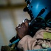 A military aviator or aircrew member is photographed in close-up profile view, wearing a flight helmet and flight suit. The individual is looking upward, with one hand raised near the chin area. The image is taken in low-light conditions with dramatic side lighting emphasizing facial features.