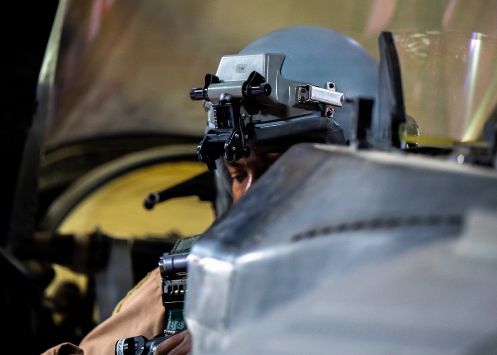 A military pilot or aircrew member is seated in what appears to be a cockpit, wearing a flight helmet equipped with night vision goggle (NVG) mounts. The individual appears to be looking through or adjusting equipment attached to the helmet. The image is taken from a rear-side angle inside or near an aircraft.