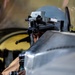 A military pilot or aircrew member is seated in what appears to be a cockpit, wearing a flight helmet equipped with night vision goggle (NVG) mounts. The individual appears to be looking through or adjusting equipment attached to the helmet. The image is taken from a rear-side angle inside or near an aircraft.