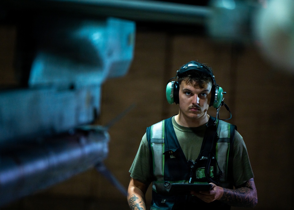 A young male technician or ground crew member is photographed in a dimly lit industrial or hangar environment. He is wearing green hearing protection headset with a communication microphone, a reflective safety vest over an olive green t-shirt, and is holding a tablet or electronic device. A blurred aircraft component is visible in the foreground, suggesting an aviation maintenance or flight deck setting.