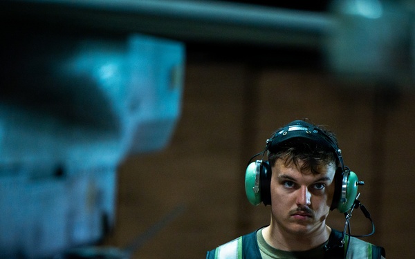 A young male technician or ground crew member is photographed in a dimly lit industrial or hangar environment. He is wearing green hearing protection headset with a communication microphone, a reflective safety vest over an olive green t-shirt, and is holding a tablet or electronic device. A blurred aircraft component is visible in the foreground, suggesting an aviation maintenance or flight deck setting.