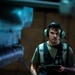 A young male technician or ground crew member is photographed in a dimly lit industrial or hangar environment. He is wearing green hearing protection headset with a communication microphone, a reflective safety vest over an olive green t-shirt, and is holding a tablet or electronic device. A blurred aircraft component is visible in the foreground, suggesting an aviation maintenance or flight deck setting.