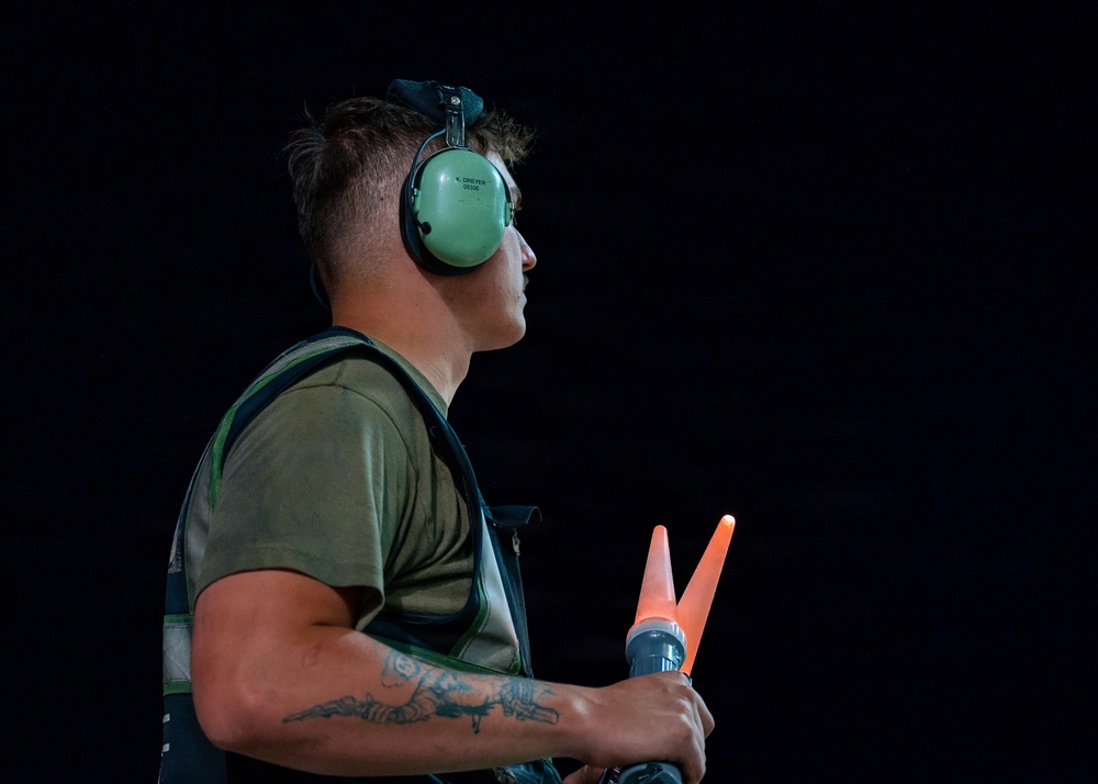 A young male aviation ground crew member is photographed in profile against a dark background, holding illuminated orange marshalling wands. He is wearing green ear protection headset and a green t-shirt with a harness or vest. The dramatic lighting isolates the subject, highlighting his focused expression and the glowing wands.