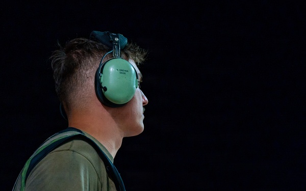 A young male aviation ground crew member is photographed in profile against a dark background, holding illuminated orange marshalling wands. He is wearing green ear protection headset and a green t-shirt with a harness or vest. The dramatic lighting isolates the subject, highlighting his focused expression and the glowing wands.