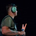 A young male aviation ground crew member is photographed in profile against a dark background, holding illuminated orange marshalling wands. He is wearing green ear protection headset and a green t-shirt with a harness or vest. The dramatic lighting isolates the subject, highlighting his focused expression and the glowing wands.