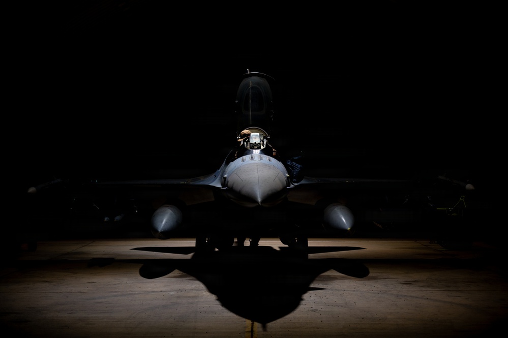 A military fighter jet is dramatically lit from above in what appears to be a darkened hangar, creating a striking chiaroscuro effect. The aircraft is photographed head-on, revealing its cockpit, fuselage, wings, and underwing stores. The low-key lighting emphasizes the aircraft's silhouette and casts a sharp shadow on the concrete floor below.