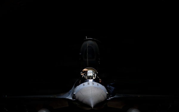 A military fighter jet is dramatically lit from above in what appears to be a darkened hangar, creating a striking chiaroscuro effect. The aircraft is photographed head-on, revealing its cockpit, fuselage, wings, and underwing stores. The low-key lighting emphasizes the aircraft's silhouette and casts a sharp shadow on the concrete floor below.