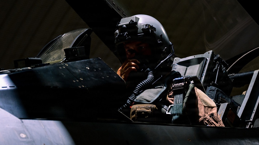 A military pilot is seated in the cockpit of a fighter aircraft, wearing a modern flight helmet equipped with night vision goggles or a helmet-mounted display system. The image is dramatically lit with strong contrast, highlighting the pilot's face and equipment against a dark background. The cockpit interior shows various instruments, panels, and tactical gear.
