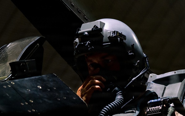 A military pilot is seated in the cockpit of a fighter aircraft, wearing a modern flight helmet equipped with night vision goggles or a helmet-mounted display system. The image is dramatically lit with strong contrast, highlighting the pilot's face and equipment against a dark background. The cockpit interior shows various instruments, panels, and tactical gear.