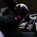 A military pilot is seated in the cockpit of a fighter aircraft, wearing a modern flight helmet equipped with night vision goggles or a helmet-mounted display system. The image is dramatically lit with strong contrast, highlighting the pilot's face and equipment against a dark background. The cockpit interior shows various instruments, panels, and tactical gear.