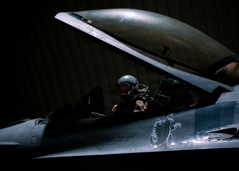 A military pilot sits in the open cockpit of a fighter jet, likely an F-16 Fighting Falcon, inside a darkened hangar. The pilot is wearing a flight helmet and flight suit, conducting what appears to be pre-flight checks or cockpit familiarization. The aircraft features a distinctive skull and dragon unit insignia painted on the fuselage.