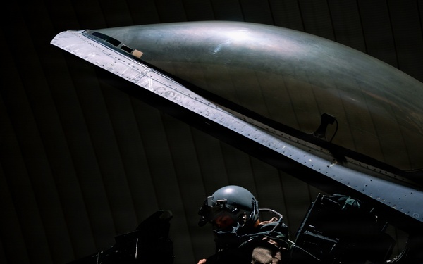 A military pilot sits in the open cockpit of a fighter jet, likely an F-16 Fighting Falcon, inside a darkened hangar. The pilot is wearing a flight helmet and flight suit, conducting what appears to be pre-flight checks or cockpit familiarization. The aircraft features a distinctive skull and dragon unit insignia painted on the fuselage.