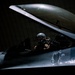 A military pilot sits in the open cockpit of a fighter jet, likely an F-16 Fighting Falcon, inside a darkened hangar. The pilot is wearing a flight helmet and flight suit, conducting what appears to be pre-flight checks or cockpit familiarization. The aircraft features a distinctive skull and dragon unit insignia painted on the fuselage.