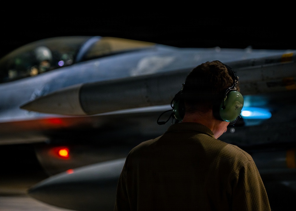 A military ground crew member wearing green hearing protection headset is observed from behind, monitoring a fighter jet aircraft during nighttime operations. The aircraft, consistent with an F-16 Fighting Falcon configuration, is illuminated against a dark background with visible navigation/warning lights active. The scene depicts a pre-flight or post-flight ground operations scenario on a military airfield.
