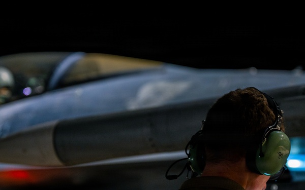 A military ground crew member wearing green hearing protection headset is observed from behind, monitoring a fighter jet aircraft during nighttime operations. The aircraft, consistent with an F-16 Fighting Falcon configuration, is illuminated against a dark background with visible navigation/warning lights active. The scene depicts a pre-flight or post-flight ground operations scenario on a military airfield.