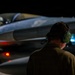 A military ground crew member wearing green hearing protection headset is observed from behind, monitoring a fighter jet aircraft during nighttime operations. The aircraft, consistent with an F-16 Fighting Falcon configuration, is illuminated against a dark background with visible navigation/warning lights active. The scene depicts a pre-flight or post-flight ground operations scenario on a military airfield.