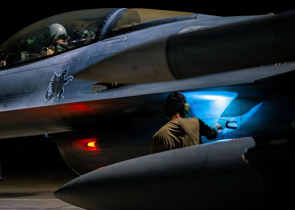 A night operations scene showing a military fighter jet, identifiable as an F-16 Fighting Falcon, being serviced on a flight line. A pilot in full flight gear sits in the cockpit while a ground crew technician inspects or services a panel on the fuselage using a bright blue light source. The dramatic low-light photography highlights the aircraft's markings, ordnance, and operational readiness activity.