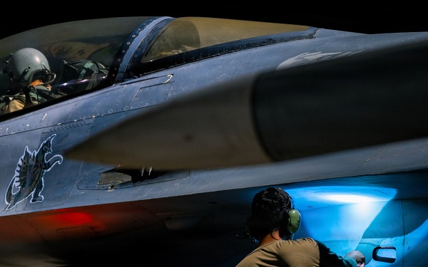 A night operations scene showing a military fighter jet, identifiable as an F-16 Fighting Falcon, being serviced on a flight line. A pilot in full flight gear sits in the cockpit while a ground crew technician inspects or services a panel on the fuselage using a bright blue light source. The dramatic low-light photography highlights the aircraft's markings, ordnance, and operational readiness activity.