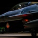 A night-time military flight line scene showing an F-16 Fighting Falcon fighter jet with tail number 894, featuring unit nose art including a bird and animal insignia. A ground crew member stands in front of the aircraft holding illuminated red marshalling wands in an X formation, wearing hearing protection. A munitions cart loaded with green ordnance is visible in the background to the left.