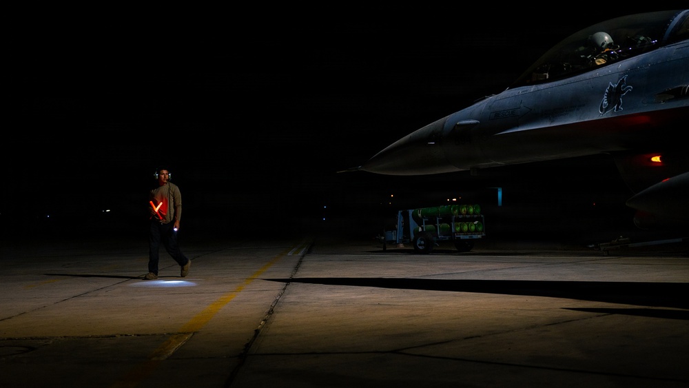 A military ground crew member stands on a darkened flight line at night, holding illuminated red marshalling wands crossed in an 'X' formation near a fighter jet. A pilot is visible in the cockpit of what appears to be an F-16 Fighting Falcon, with a munitions cart loaded with green ordnance visible in the background. The scene depicts a nighttime pre-flight or taxi operation at a military airbase.