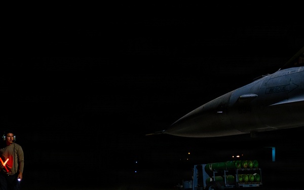 A military ground crew member stands on a darkened flight line at night, holding illuminated red marshalling wands crossed in an 'X' formation near a fighter jet. A pilot is visible in the cockpit of what appears to be an F-16 Fighting Falcon, with a munitions cart loaded with green ordnance visible in the background. The scene depicts a nighttime pre-flight or taxi operation at a military airbase.