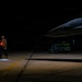 A military ground crew member stands on a darkened flight line at night, holding illuminated red marshalling wands crossed in an 'X' formation near a fighter jet. A pilot is visible in the cockpit of what appears to be an F-16 Fighting Falcon, with a munitions cart loaded with green ordnance visible in the background. The scene depicts a nighttime pre-flight or taxi operation at a military airbase.