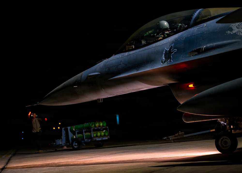 A military jet fighter, identifiable as an F-16 Fighting Falcon, is photographed on a flight line at night with a pilot seated in the cockpit. Ground crew personnel are visible in the background, and a munitions cart loaded with green ordnance is parked nearby. The aircraft displays a unit insignia (appears to be a griffin or dragon emblem) on its fuselage and has navigation/position lights illuminated.