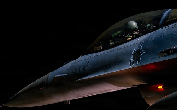 A military jet fighter, identifiable as an F-16 Fighting Falcon, is photographed on a flight line at night with a pilot seated in the cockpit. Ground crew personnel are visible in the background, and a munitions cart loaded with green ordnance is parked nearby. The aircraft displays a unit insignia (appears to be a griffin or dragon emblem) on its fuselage and has navigation/position lights illuminated.