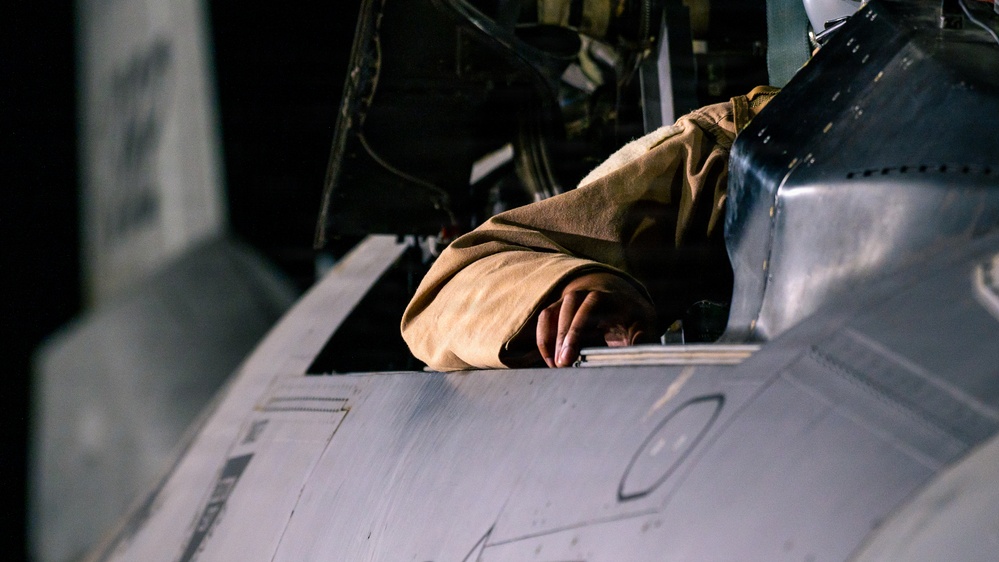 A person wearing a tan/khaki flight suit or jacket is leaning into or over the cockpit area of a military aircraft. The image is taken at close range, focusing on the upper fuselage and cockpit rim of what appears to be a jet aircraft. The dark background suggests this was taken in a hangar or during nighttime operations.