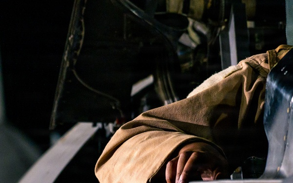 A person wearing a tan/khaki flight suit or jacket is leaning into or over the cockpit area of a military aircraft. The image is taken at close range, focusing on the upper fuselage and cockpit rim of what appears to be a jet aircraft. The dark background suggests this was taken in a hangar or during nighttime operations.
