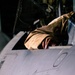 A person wearing a tan/khaki flight suit or jacket is leaning into or over the cockpit area of a military aircraft. The image is taken at close range, focusing on the upper fuselage and cockpit rim of what appears to be a jet aircraft. The dark background suggests this was taken in a hangar or during nighttime operations.