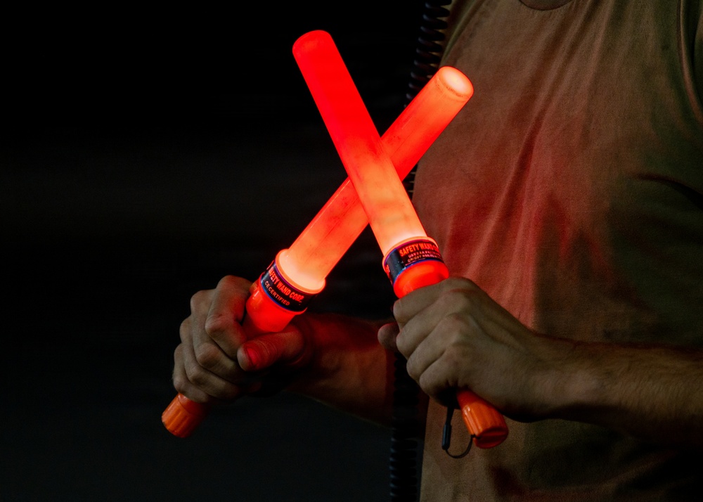 A person is holding two illuminated red safety wands crossed in an X formation against a dark background. The wands are battery-powered light batons manufactured by Safety Wand Corp, commonly used for traffic control, aircraft marshaling, or emergency signaling. The red glow from the wands illuminates the person's hands and torso.