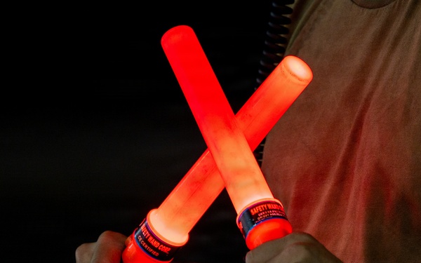 A person is holding two illuminated red safety wands crossed in an X formation against a dark background. The wands are battery-powered light batons manufactured by Safety Wand Corp, commonly used for traffic control, aircraft marshaling, or emergency signaling. The red glow from the wands illuminates the person's hands and torso.