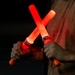 A person is holding two illuminated red safety wands crossed in an X formation against a dark background. The wands are battery-powered light batons manufactured by Safety Wand Corp, commonly used for traffic control, aircraft marshaling, or emergency signaling. The red glow from the wands illuminates the person's hands and torso.
