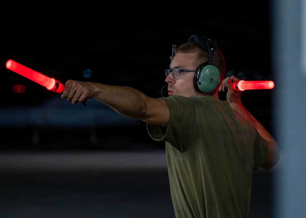 A young male aircraft marshaller is directing aircraft at night using two illuminated red signal wands. He is wearing green aviation ear protection headsets, glasses, and an olive/green t-shirt. The dark background suggests a nighttime airfield or flight line operation.