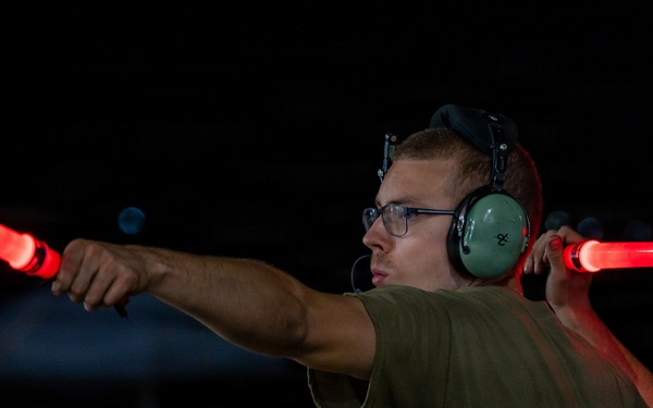 A young male aircraft marshaller is directing aircraft at night using two illuminated red signal wands. He is wearing green aviation ear protection headsets, glasses, and an olive/green t-shirt. The dark background suggests a nighttime airfield or flight line operation.