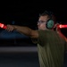 A young male aircraft marshaller is directing aircraft at night using two illuminated red signal wands. He is wearing green aviation ear protection headsets, glasses, and an olive/green t-shirt. The dark background suggests a nighttime airfield or flight line operation.