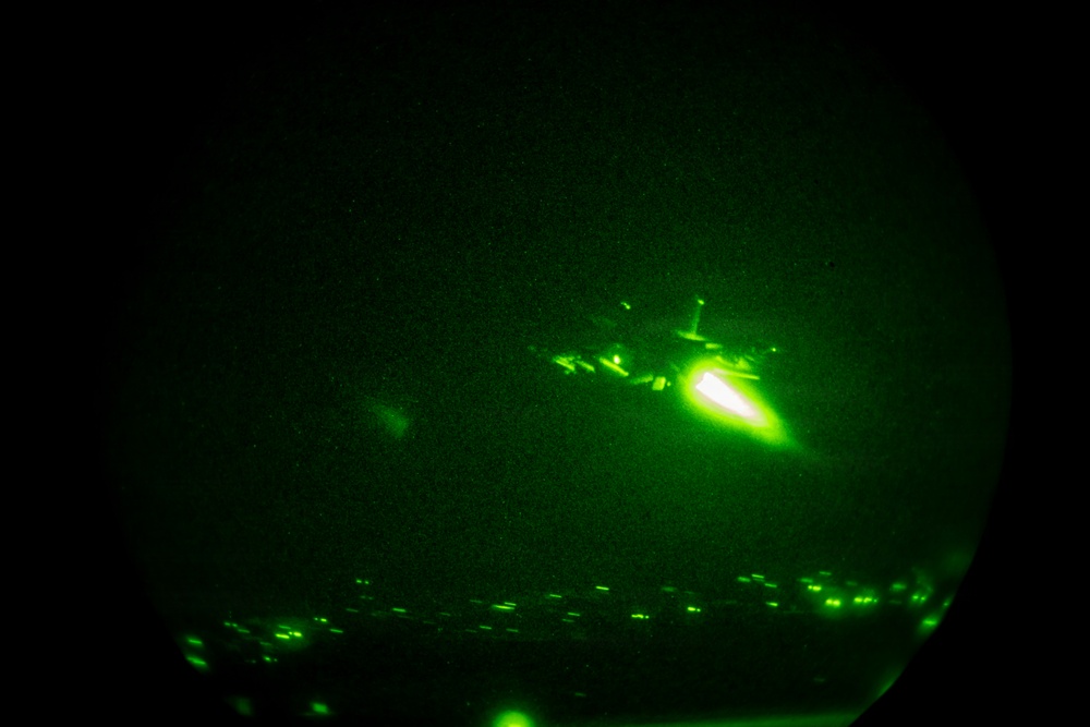 A night vision image captured through a circular lens showing a military aircraft in flight with a bright engine exhaust or flare visible. The image is taken through night vision optics, rendering everything in characteristic green monochrome tones. Lights consistent with a populated area or airfield are visible along the bottom edge of the frame.