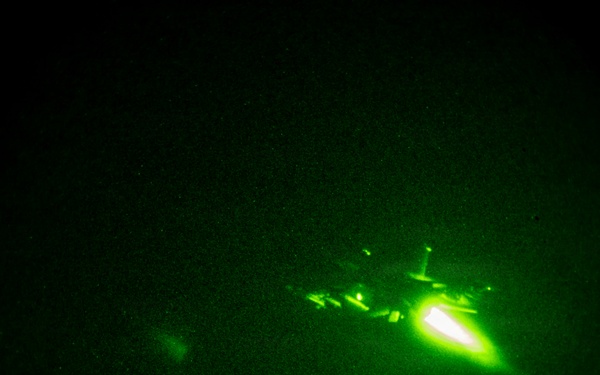 A night vision image captured through a circular lens showing a military aircraft in flight with a bright engine exhaust or flare visible. The image is taken through night vision optics, rendering everything in characteristic green monochrome tones. Lights consistent with a populated area or airfield are visible along the bottom edge of the frame.