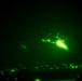 A night vision image captured through a circular lens showing a military aircraft in flight with a bright engine exhaust or flare visible. The image is taken through night vision optics, rendering everything in characteristic green monochrome tones. Lights consistent with a populated area or airfield are visible along the bottom edge of the frame.