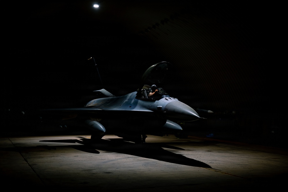 A single-seat fighter jet, consistent with an F-16 Fighting Falcon, is parked inside a darkened hangar or hardened aircraft shelter, illuminated by a single overhead spotlight. A helmeted individual is visible in the open cockpit, appearing to conduct pre-flight checks or cockpit familiarization. The dramatic low-key lighting creates strong contrast between the illuminated aircraft and the surrounding darkness.