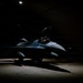 A single-seat fighter jet, consistent with an F-16 Fighting Falcon, is parked inside a darkened hangar or hardened aircraft shelter, illuminated by a single overhead spotlight. A helmeted individual is visible in the open cockpit, appearing to conduct pre-flight checks or cockpit familiarization. The dramatic low-key lighting creates strong contrast between the illuminated aircraft and the surrounding darkness.