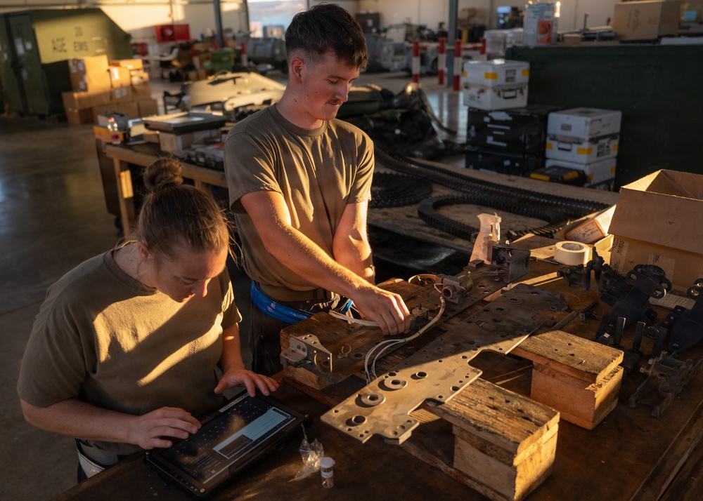 Two military personnel in olive drab t-shirts are working at a large workbench in what appears to be a maintenance or repair facility. One person is seated and operating a tablet/laptop computer while the other stands and handles mechanical components. The workspace is cluttered with various metal parts, tools, wooden blocks, and supplies in a warehouse-like environment.