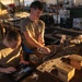 Two military personnel in olive drab t-shirts are working at a large workbench in what appears to be a maintenance or repair facility. One person is seated and operating a tablet/laptop computer while the other stands and handles mechanical components. The workspace is cluttered with various metal parts, tools, wooden blocks, and supplies in a warehouse-like environment.
