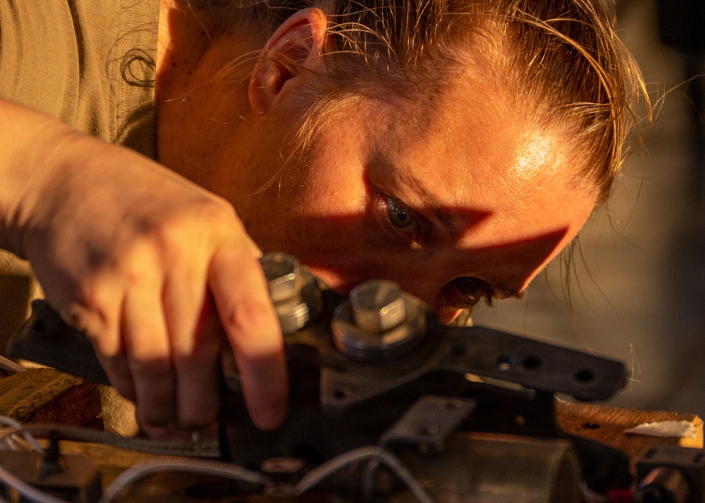 A person is closely inspecting or working on a mechanical component, leaning in with intense focus. The image is bathed in warm golden-hour light, creating dramatic shadows across the subject's face. The individual appears to be a technician or mechanic performing detailed hands-on work.